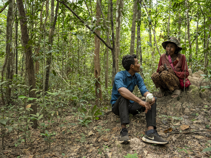 A young person in Cambodia engaging with an elderly community member while seated together in a forest.