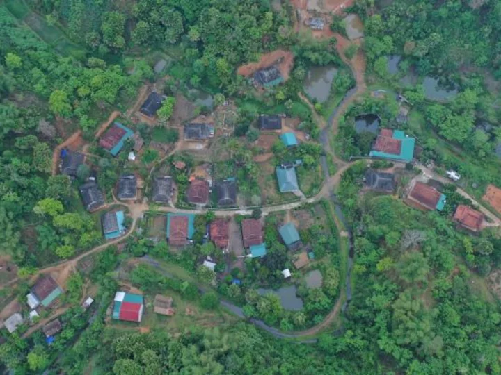 An aerial view of a rural village surrounded by dense greenery, featuring houses with colorful roofs connected by winding roads.