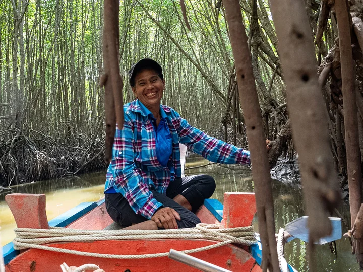 A woman in a plaid shirt sits on a small boat navigating through a mangrove forest, surrounded by roots and calm water.