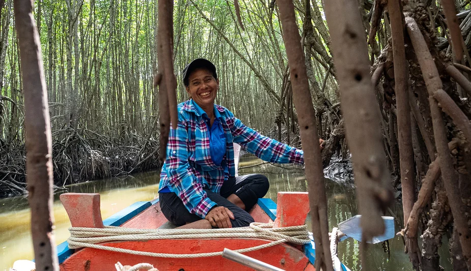 A woman in a plaid shirt sits on a small boat navigating through a mangrove forest, surrounded by roots and calm water.
