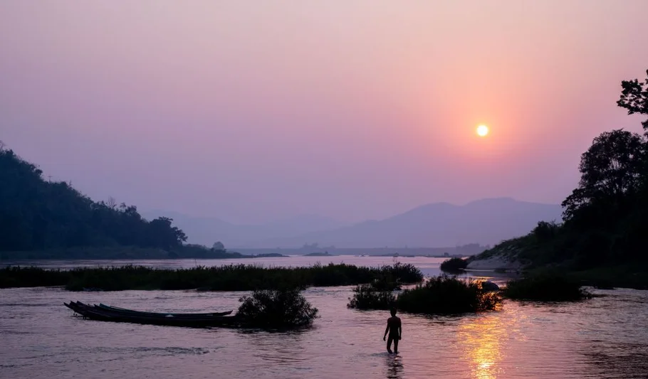 A river landscape at sunset, with a person standing in shallow water surrounded by small islands of vegetation.